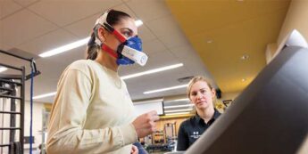 Fit student wearing breathing device gives thumbs up to another student while walking on a treadmill