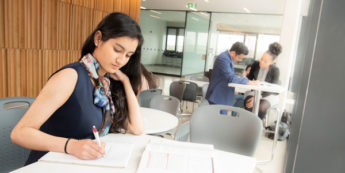 Student wearing business attire intently writing in notebook while others work in the background