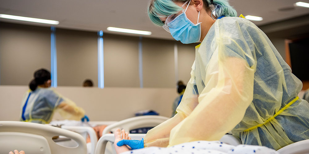 Woman wearing PPE attends to a nursing mannequin in a hospital bed