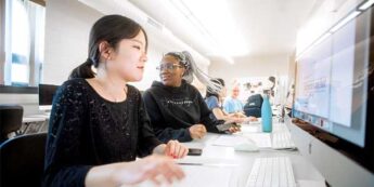 Two female students in the graphic design lab with one student working on a computer while the other looks away.