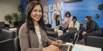 Female student smiles looking at the camera while working on a laptop in the foreground, and a group of four students sit together in the background, collaborating over another laptop.