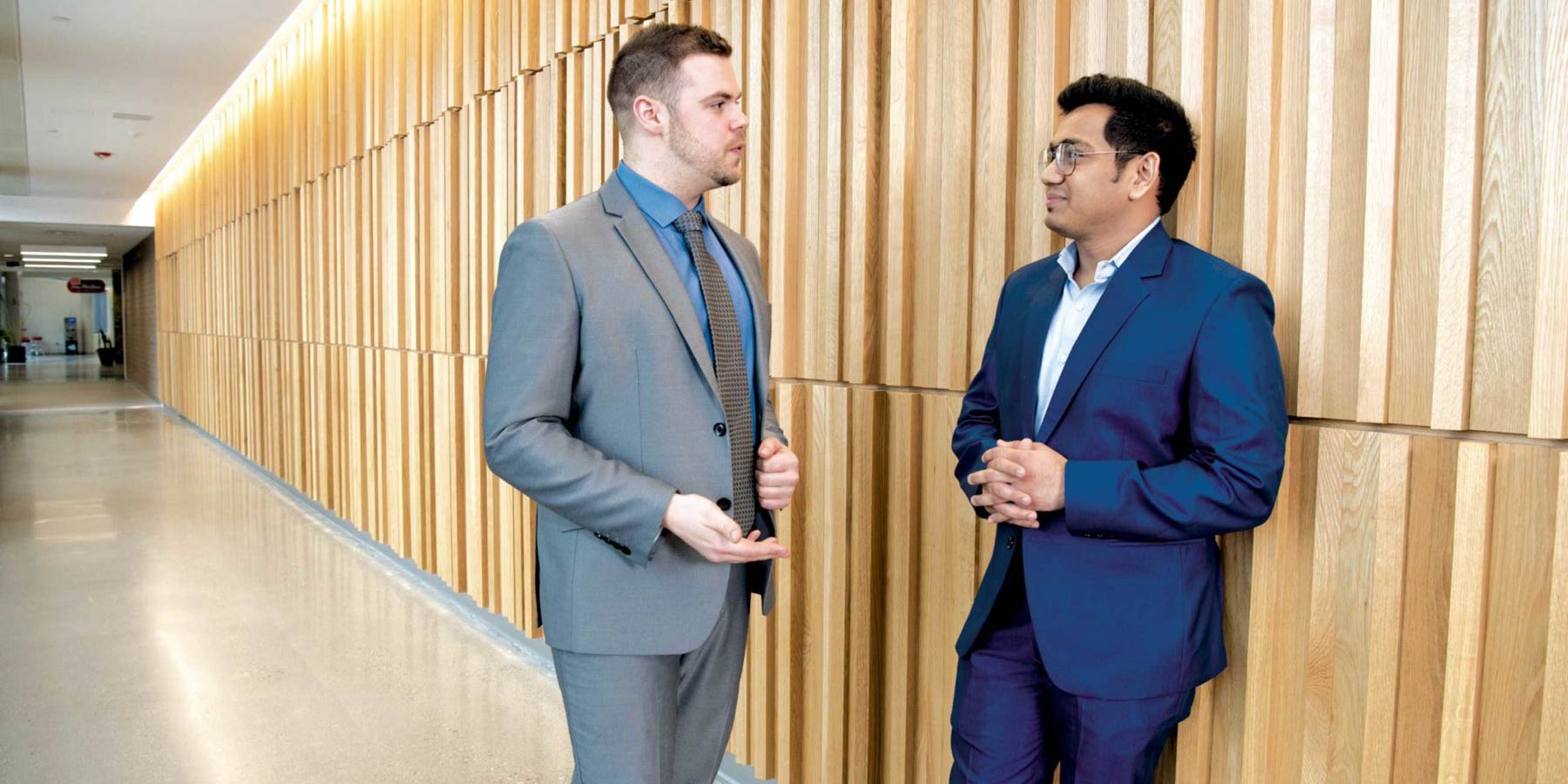 Two students in business suits chat in a modern, wood-clad hallway