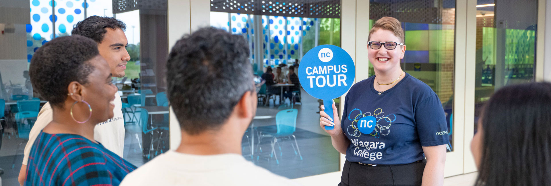 Tour guide holds up a "Campus Tour" sign while she guides visitors around the Welland campus