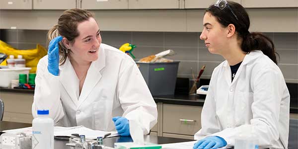 Two students in lab coats and blue gloves converse at a laboratory table with notebooks and lab equipment
