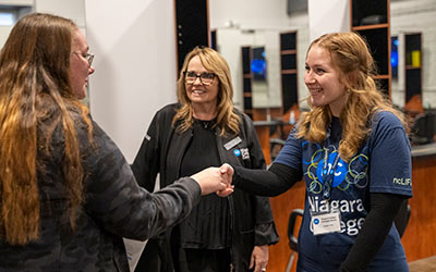 A prospective female student and current student who is working open house shake hands while a faculty member looks on.