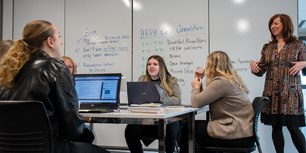 A group of students collaborates around a table while a student presents in a boardroom.