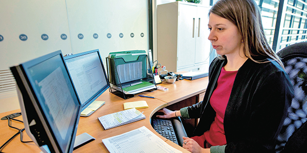 Women at desk with multiple monitors and papers in front of her.