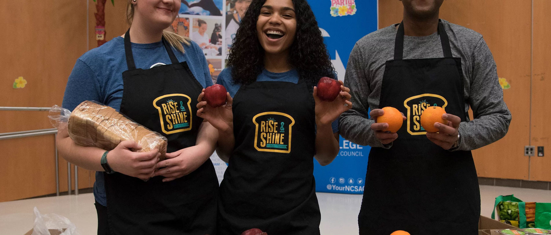 3 students holding bread and fruit wearing Rise & Shine aprons