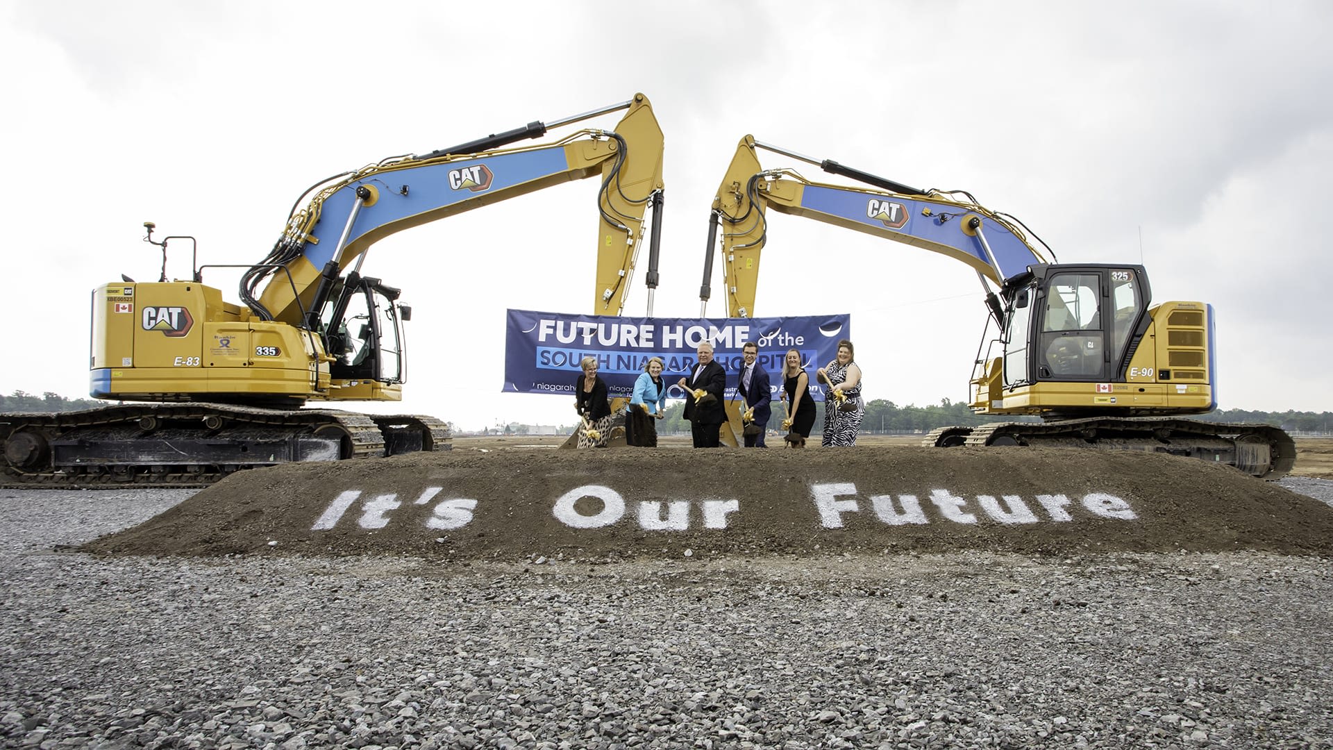 Two excavators positioned behind a sign that reads "It's Our Future" as people with shovels do a ground breaking ceremony at the new South Niagara hospital in Niagara Falls.