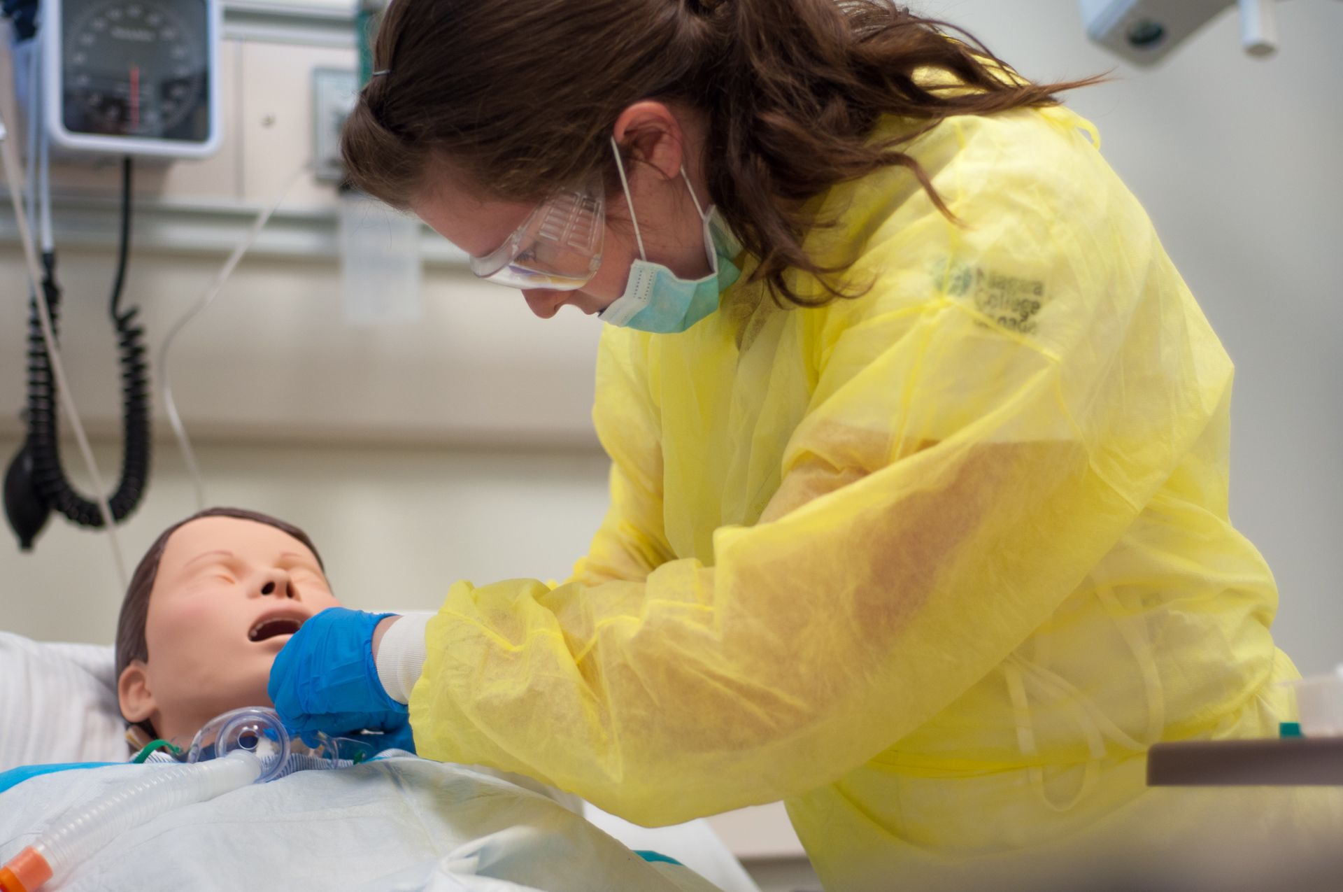 A female student wearing a yellow gown and face mask practices chest compressions on a medical mannequins.