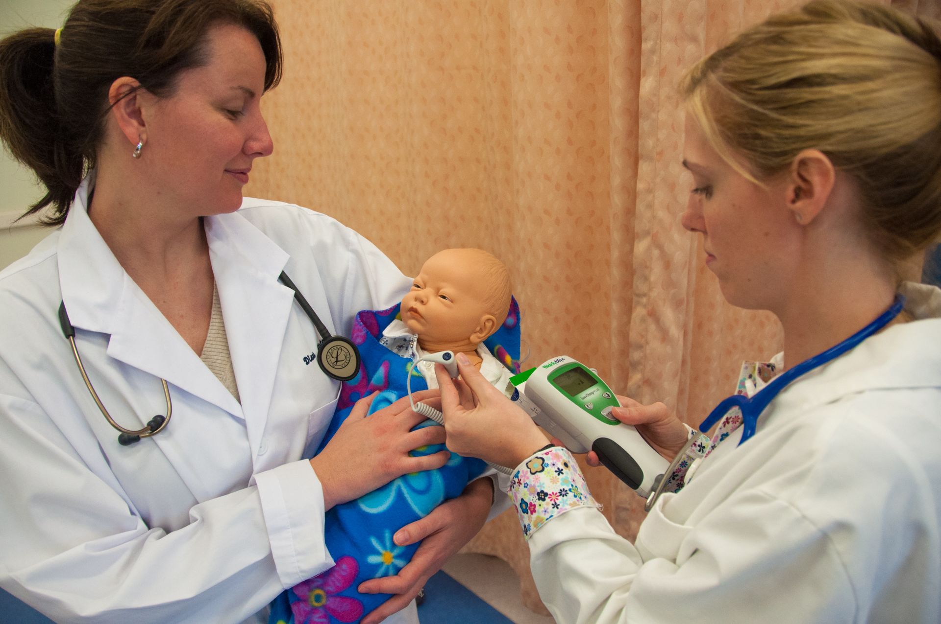  Two nursing students wearing lab coats simulate taking a baby's temperature. 