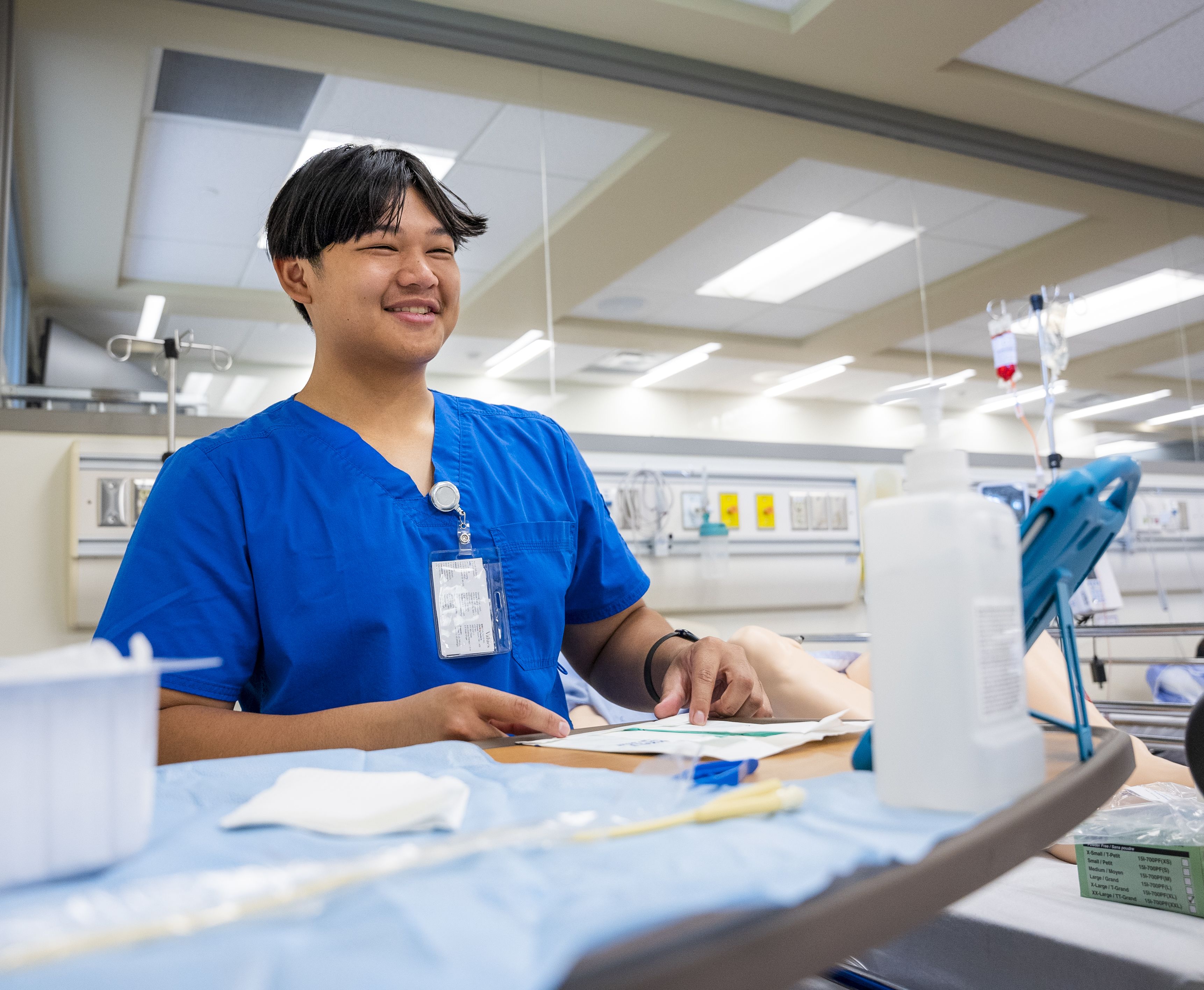 Male nursing student wearing blue Niagara College scrubs smiles inside a classroom lab.