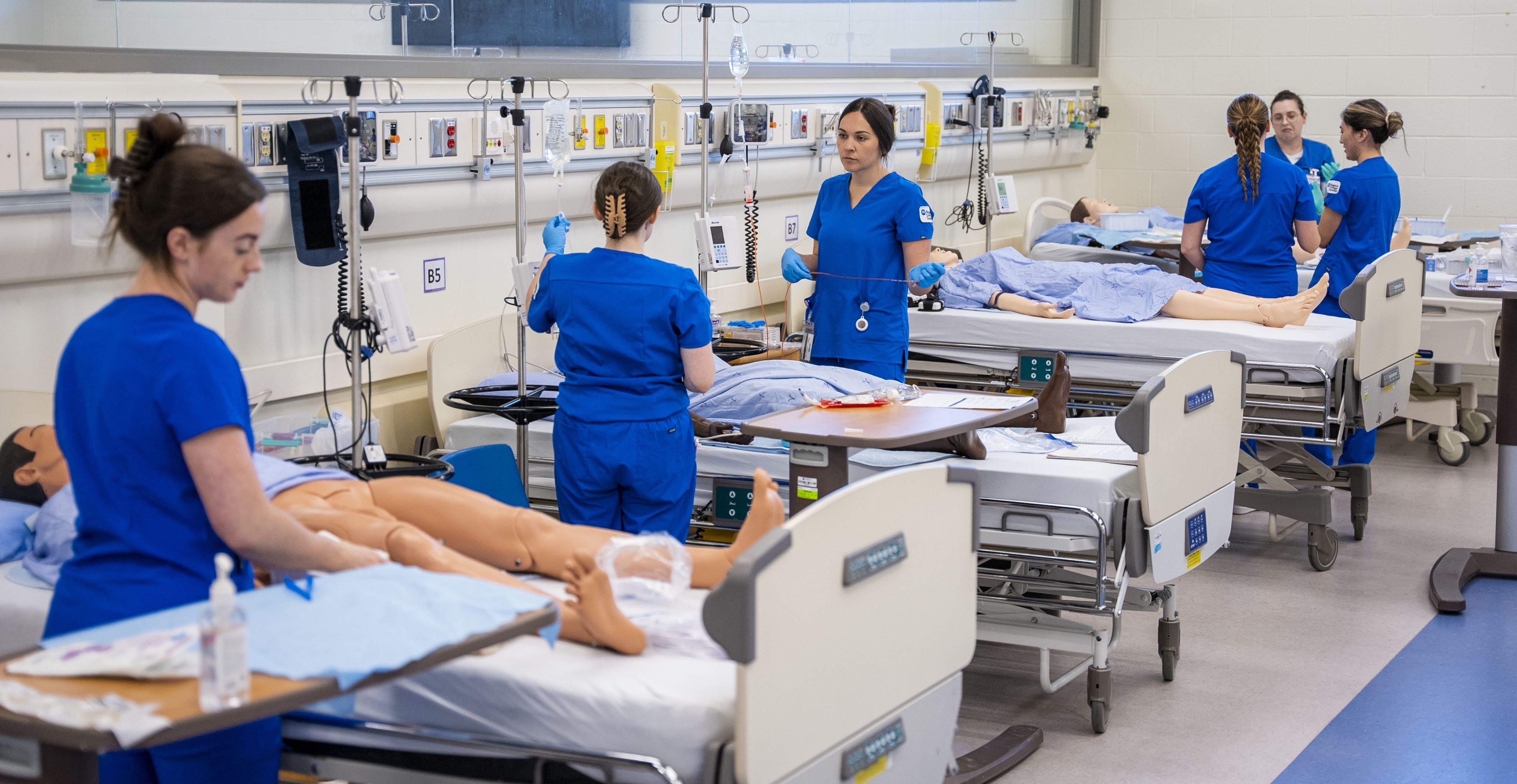 Nursing students wearing blue Niagara College scrubs in a hospital room lab doing patient checks.