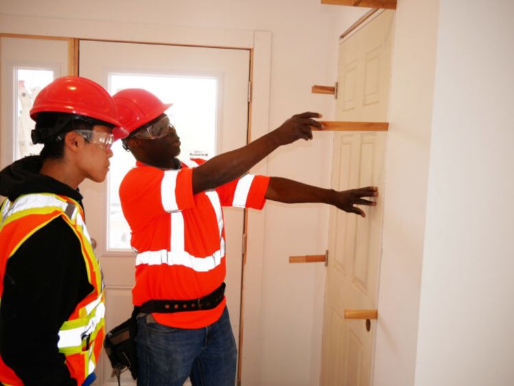 Two male students in hard hats and safety vests inspect measurements on a doorframe.