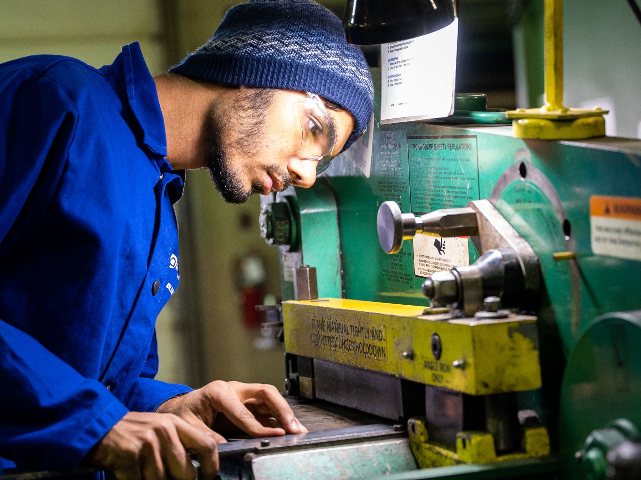 A male student wearing safety glasses, a beanie, and blue coveralls operates a green and yellow industrial machine in a workshop.