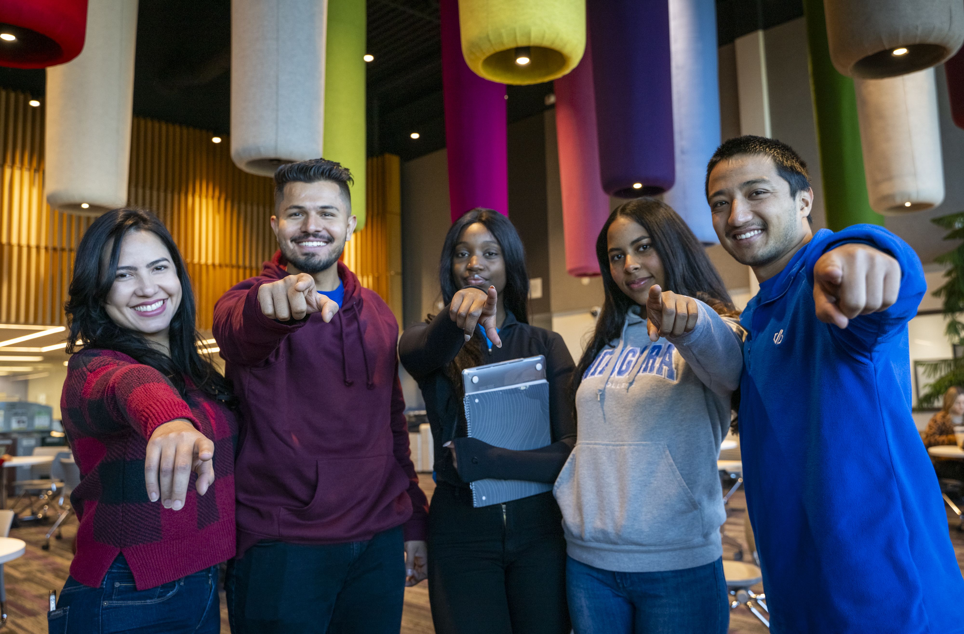 Diverse group of five students smiling and pointing their index finder at the camera.