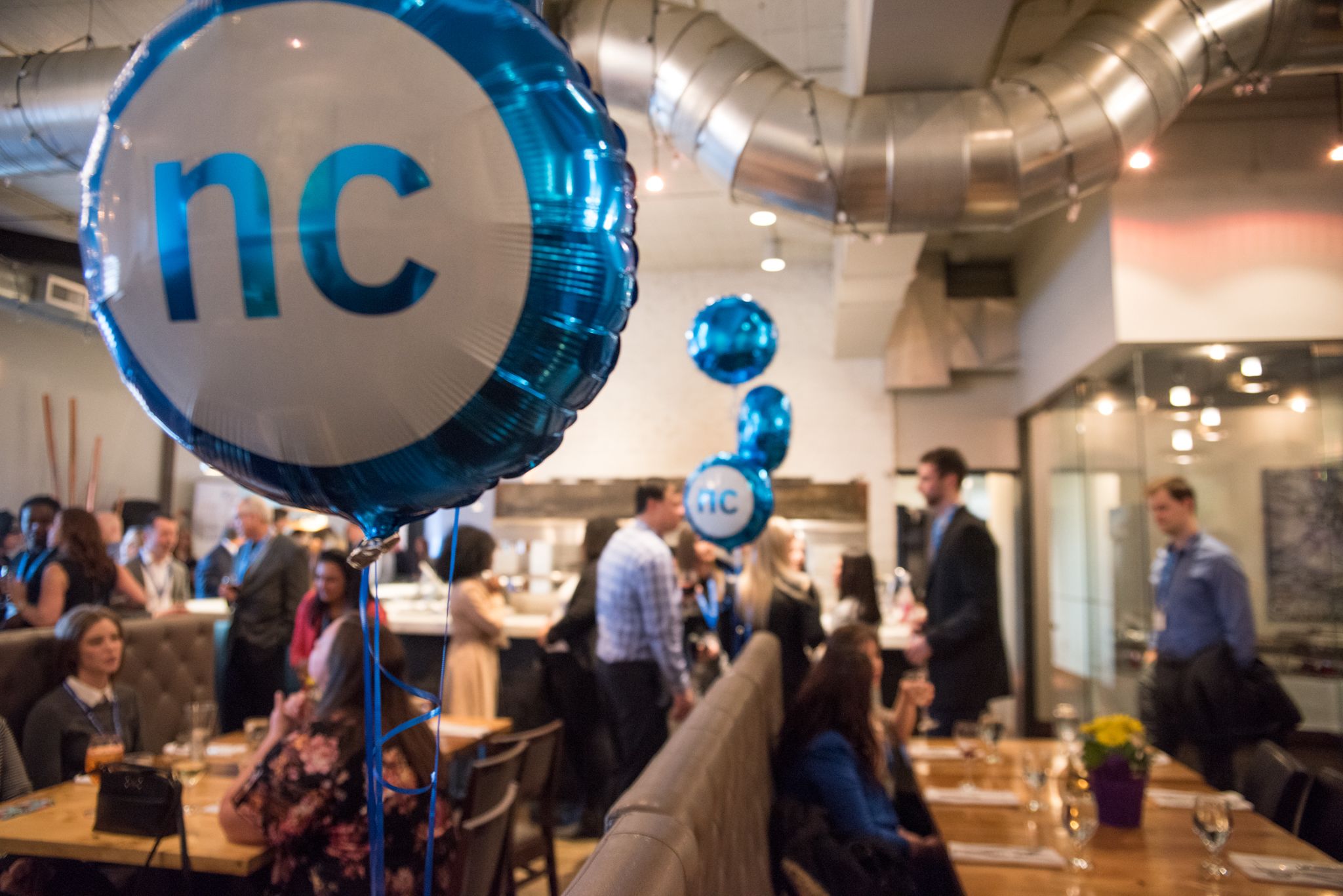 A colourful, circular NC branded balloon in the foreground with several people standing in the background and networking. 