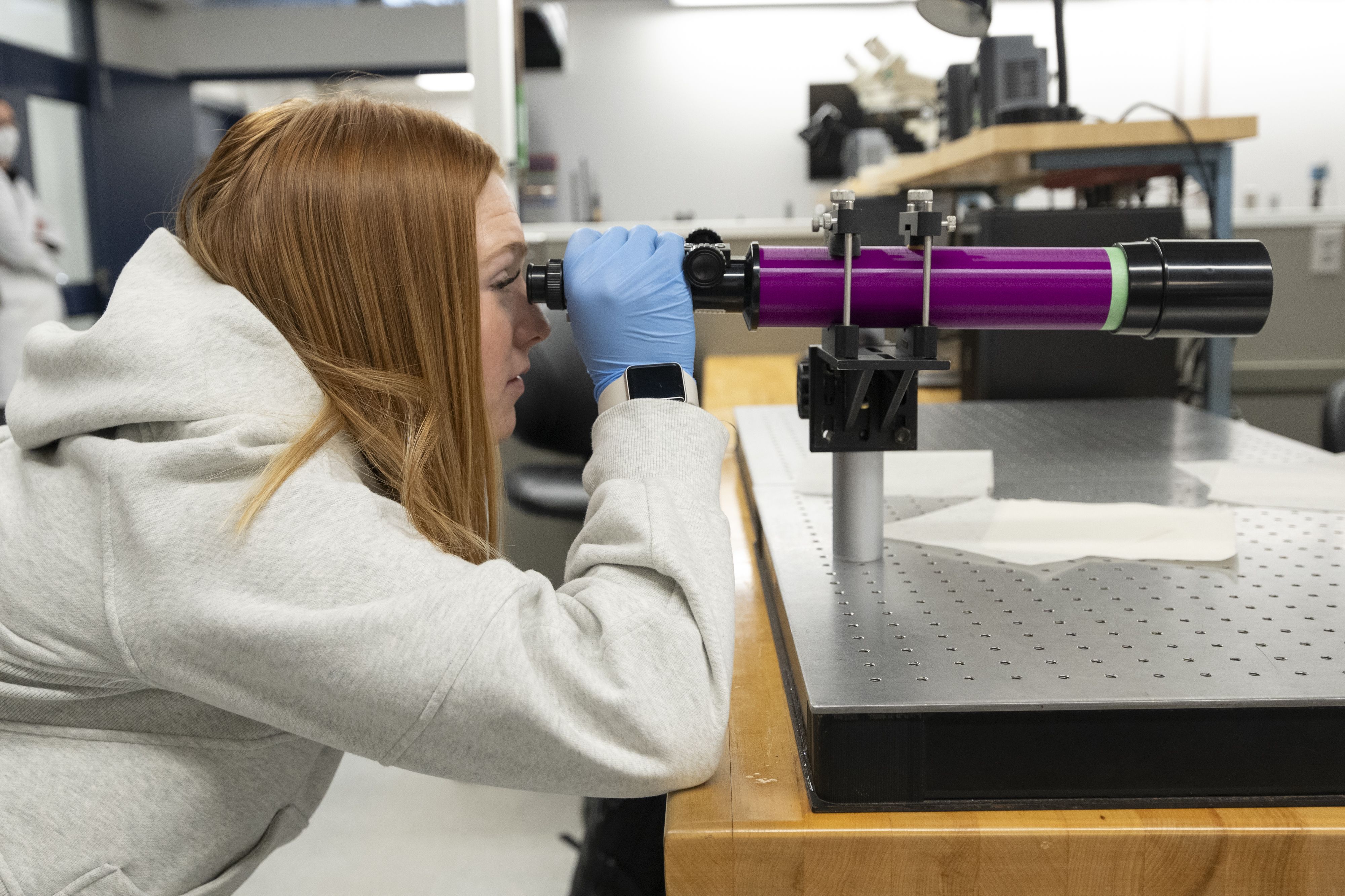 A female student wearing a white lab coat looks through a microscope in a laboratory setting.
