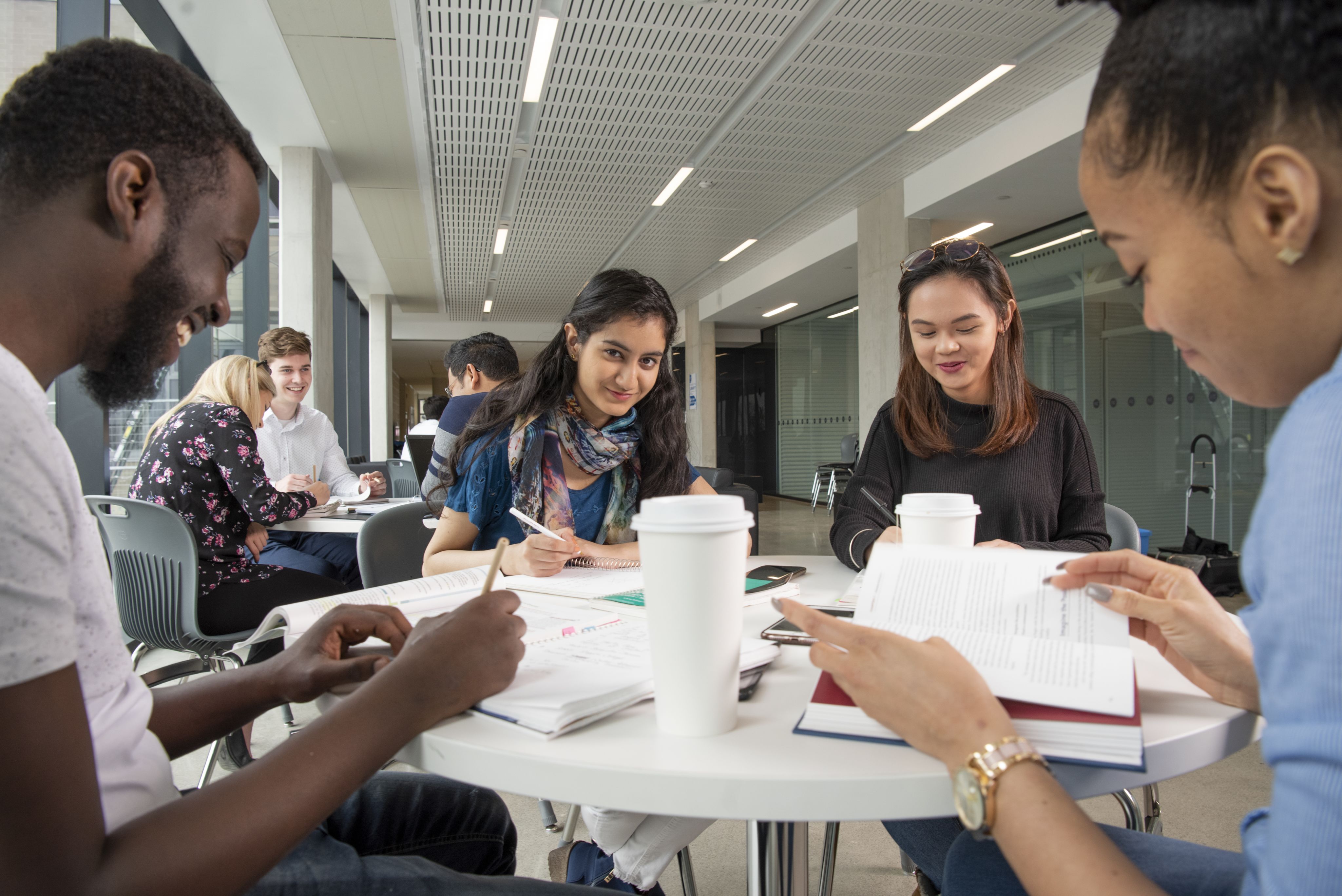 A diverse group of students study together around a table with books, papers, and coffee cups.