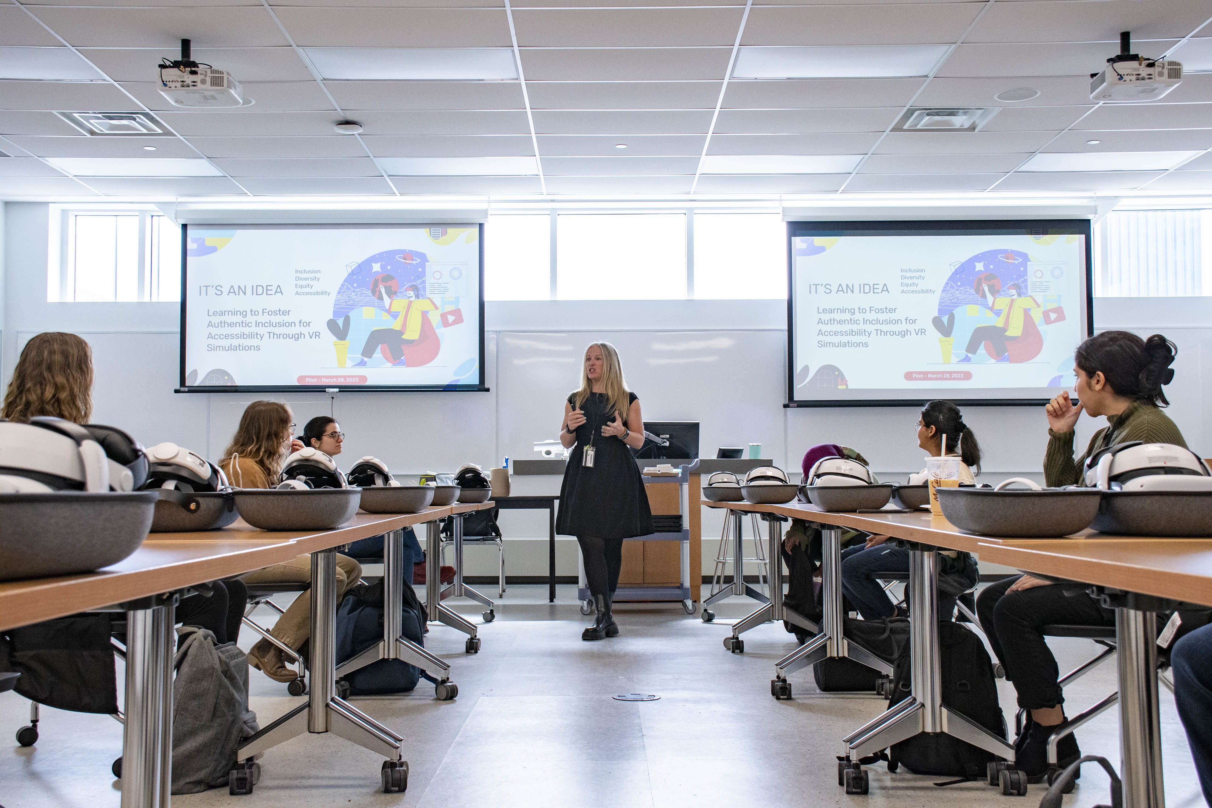 A female professor stands at the front of a classroom, delivering a presentation to engaged students seated at desks.