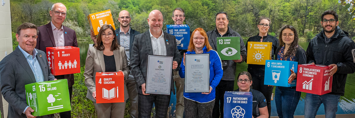 Group of NC staff poses with 2 large certificates for the UN Sustainable Development Goals signing pledge