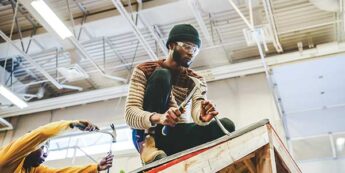 Two male students hammering on the roof of a wooden structure in the construction lab with one student in the middle of the image.