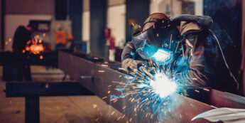 Student wearing welding mask and gloves welding a metal beam.