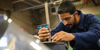 An industrial millwright student wearing safety glasses carefully measures equipment using a combination square in a workshop setting.