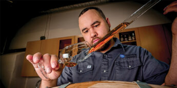 Student pouring spirit into glass over wooden barrel.