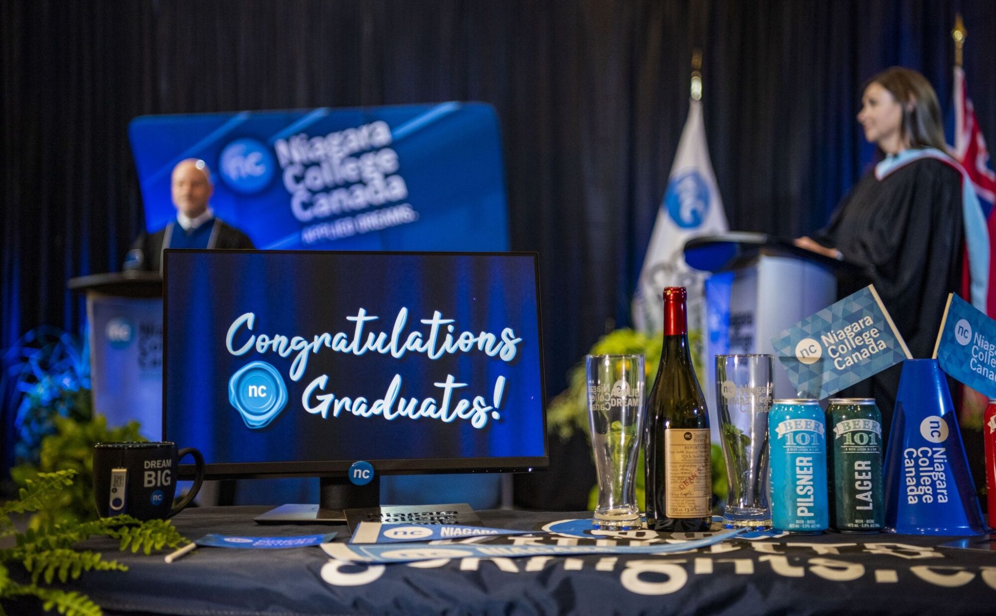 President Kennedy and Fiona Allan on the convocation stage with NC swag in the foreground, congratulations graduates on a monitor
