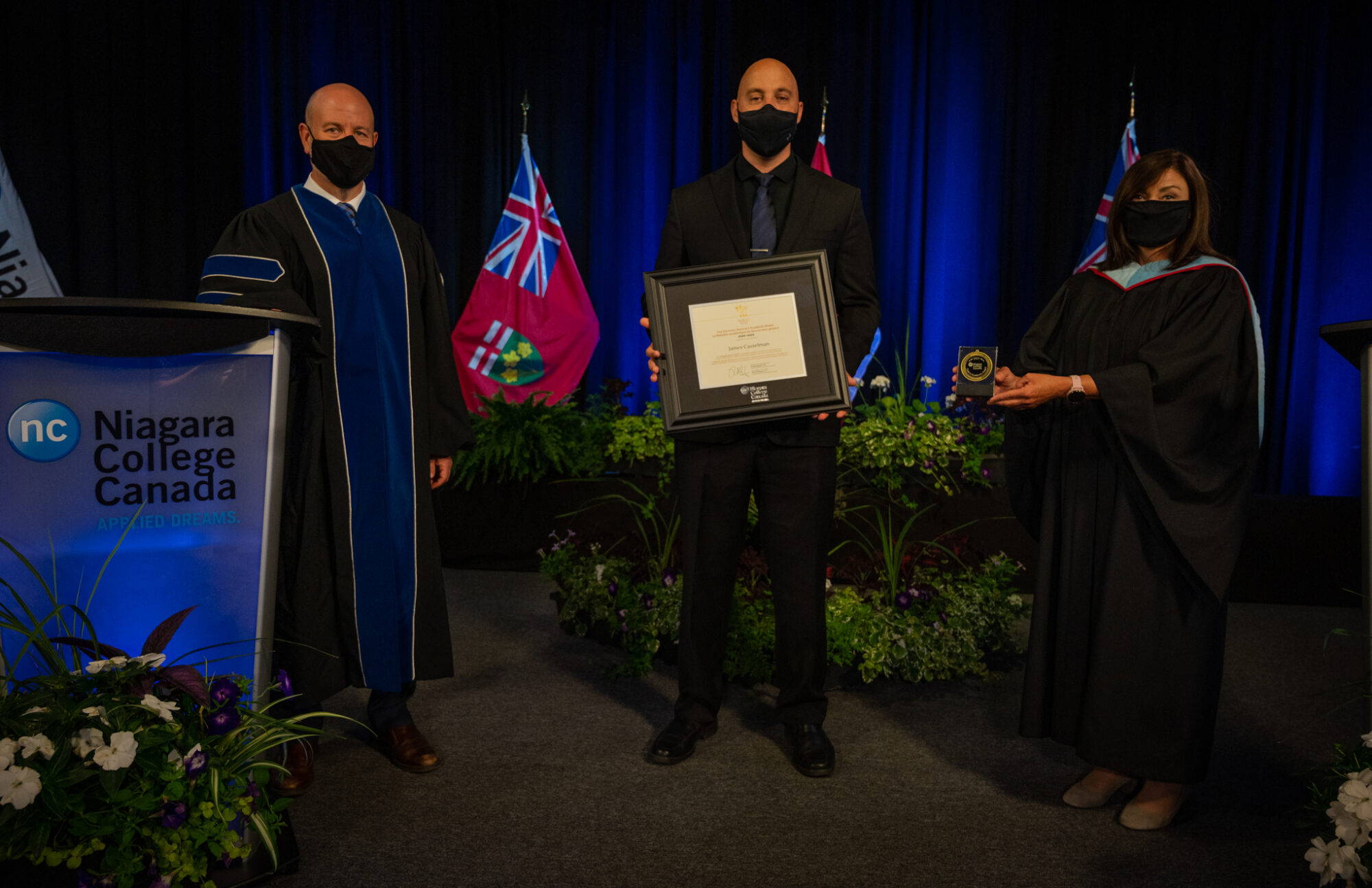 A man and woman dressed in graduation robes present a man standing between them with a framed certificate and medal.