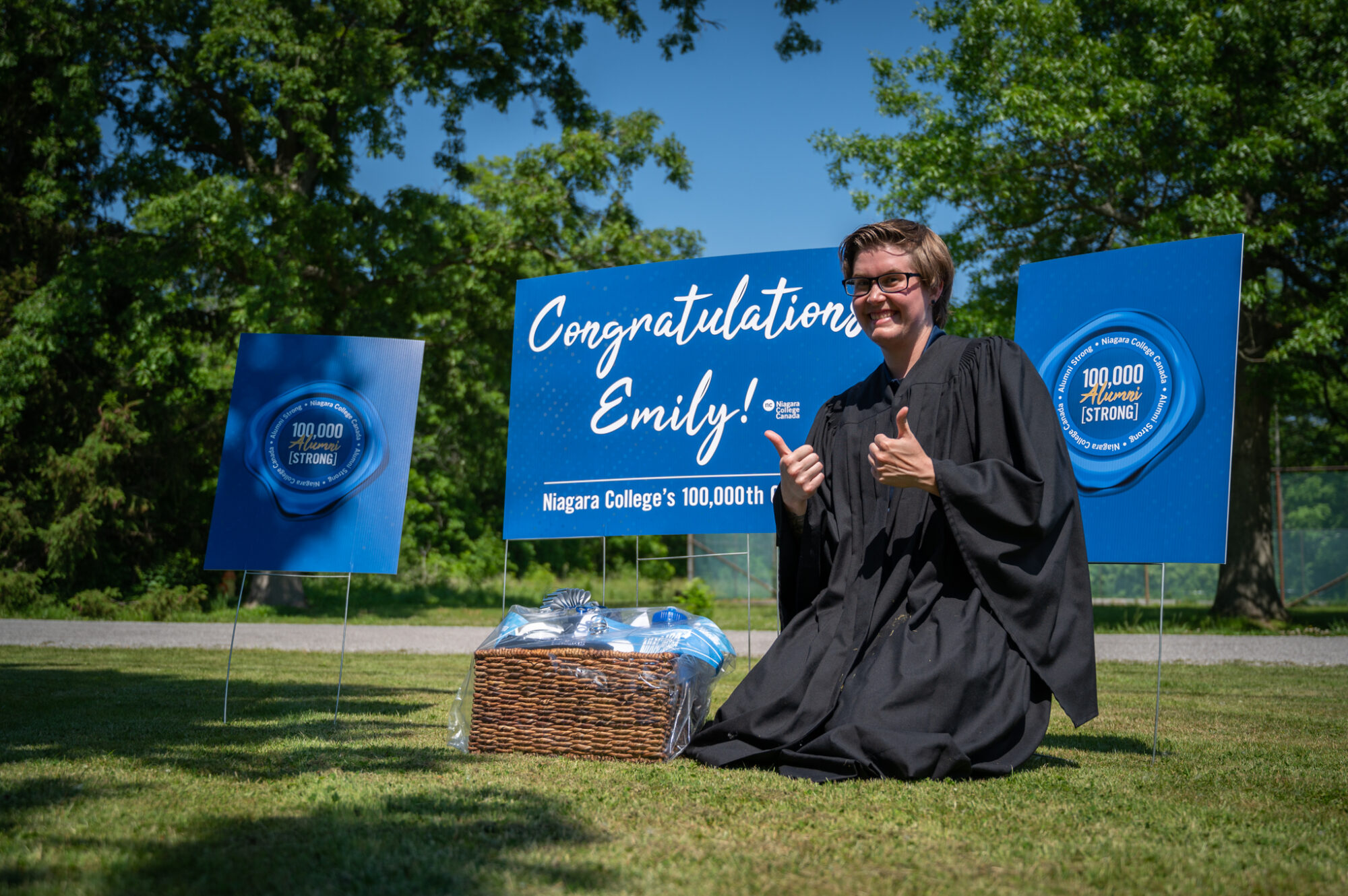 A woman in a grad gown gives the thumbs up while kneeling in front of a sign that says she's the 100,00th graduate.