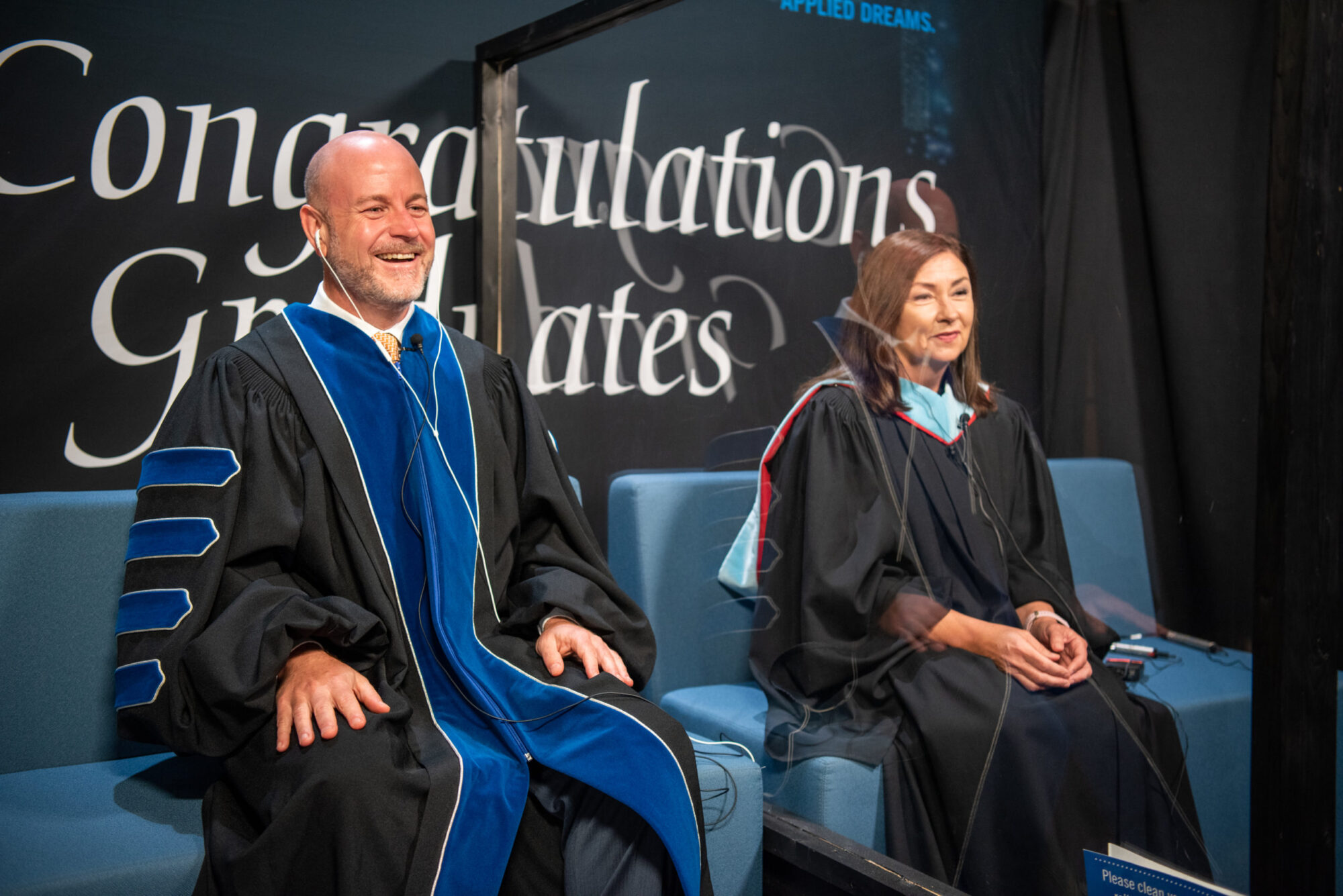 A man and woman wearing graduation regalia speak to a camera.
