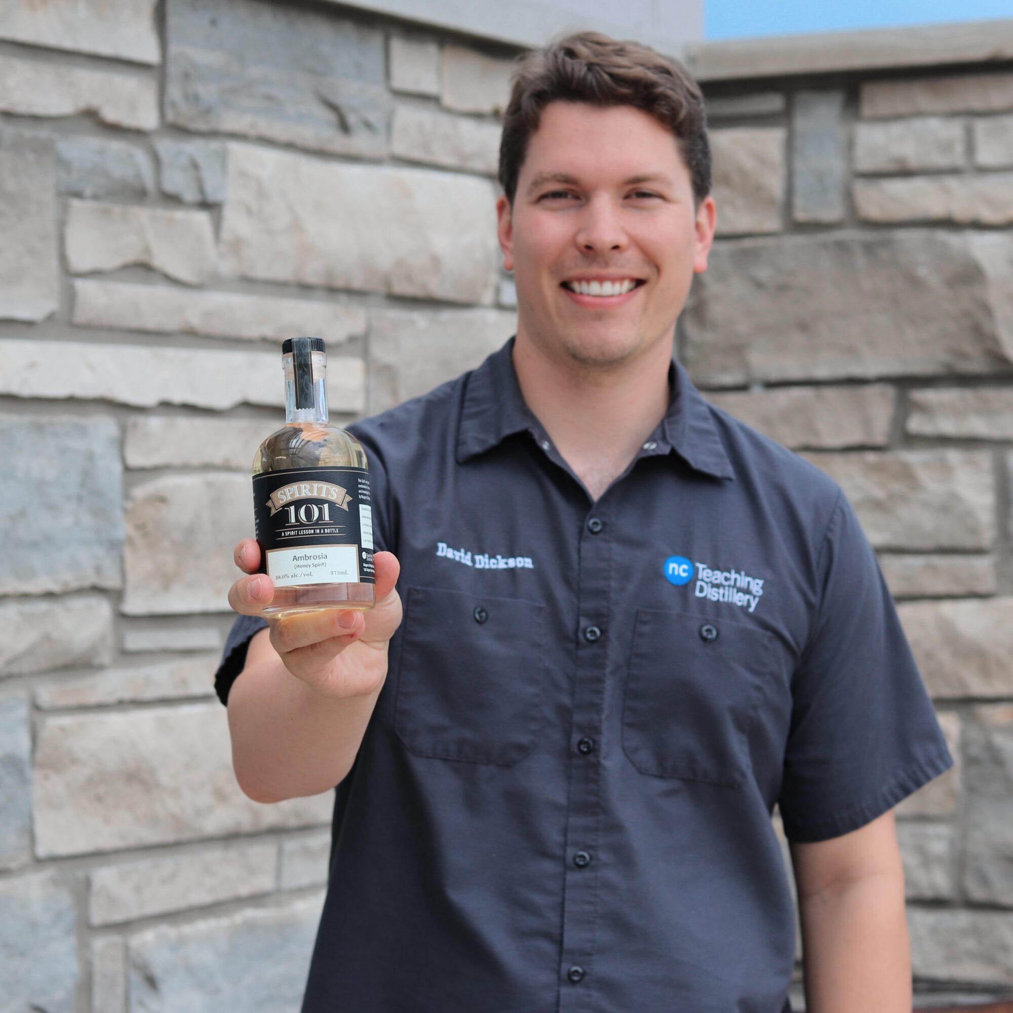 A man standing against a brick wall smiles and holds a small glass bottle filled with a spirit called Ambrosia