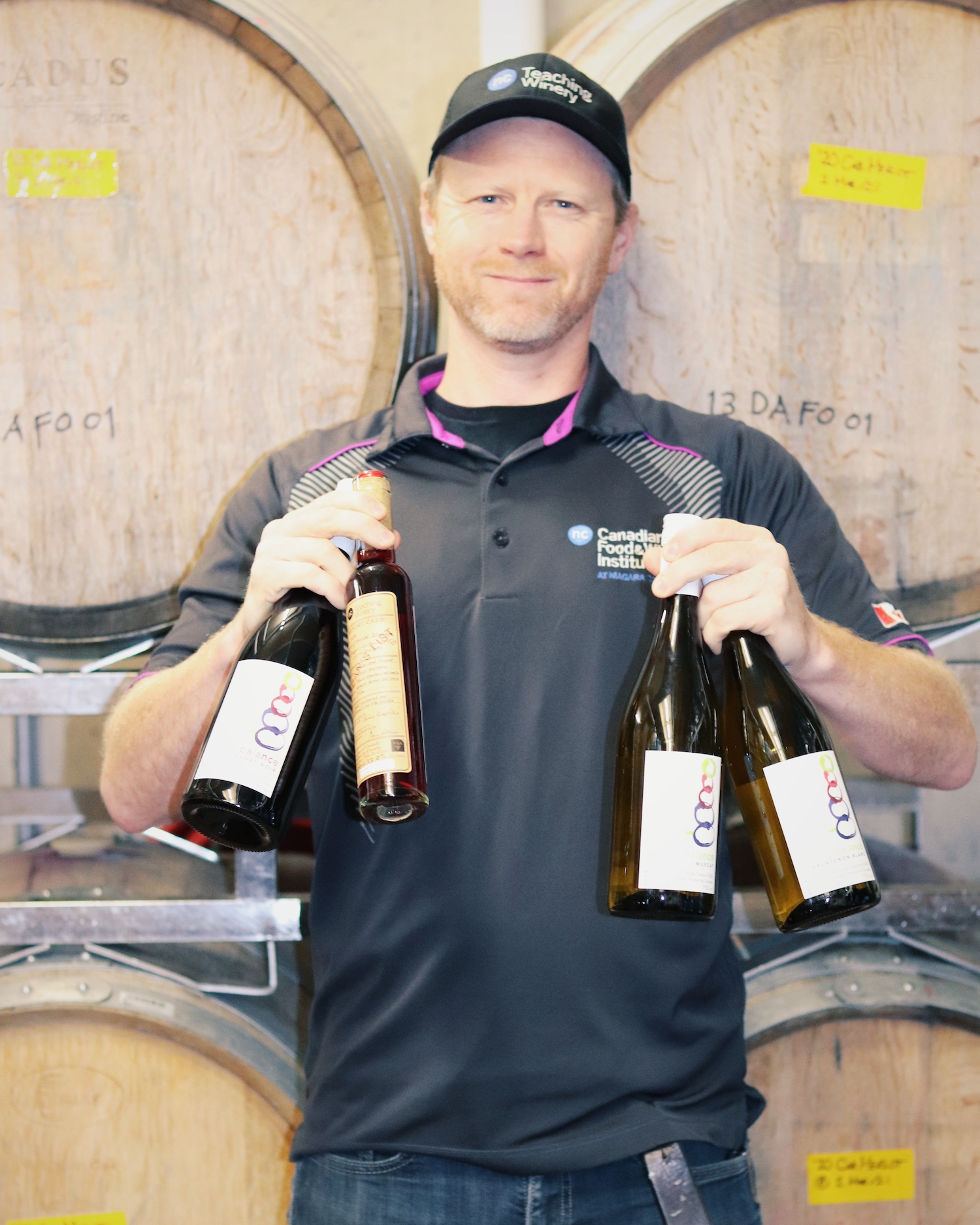 A man wearing a baseball cap stands in front of wine barrels holding four bottles of wine in his hands. He is smiling.