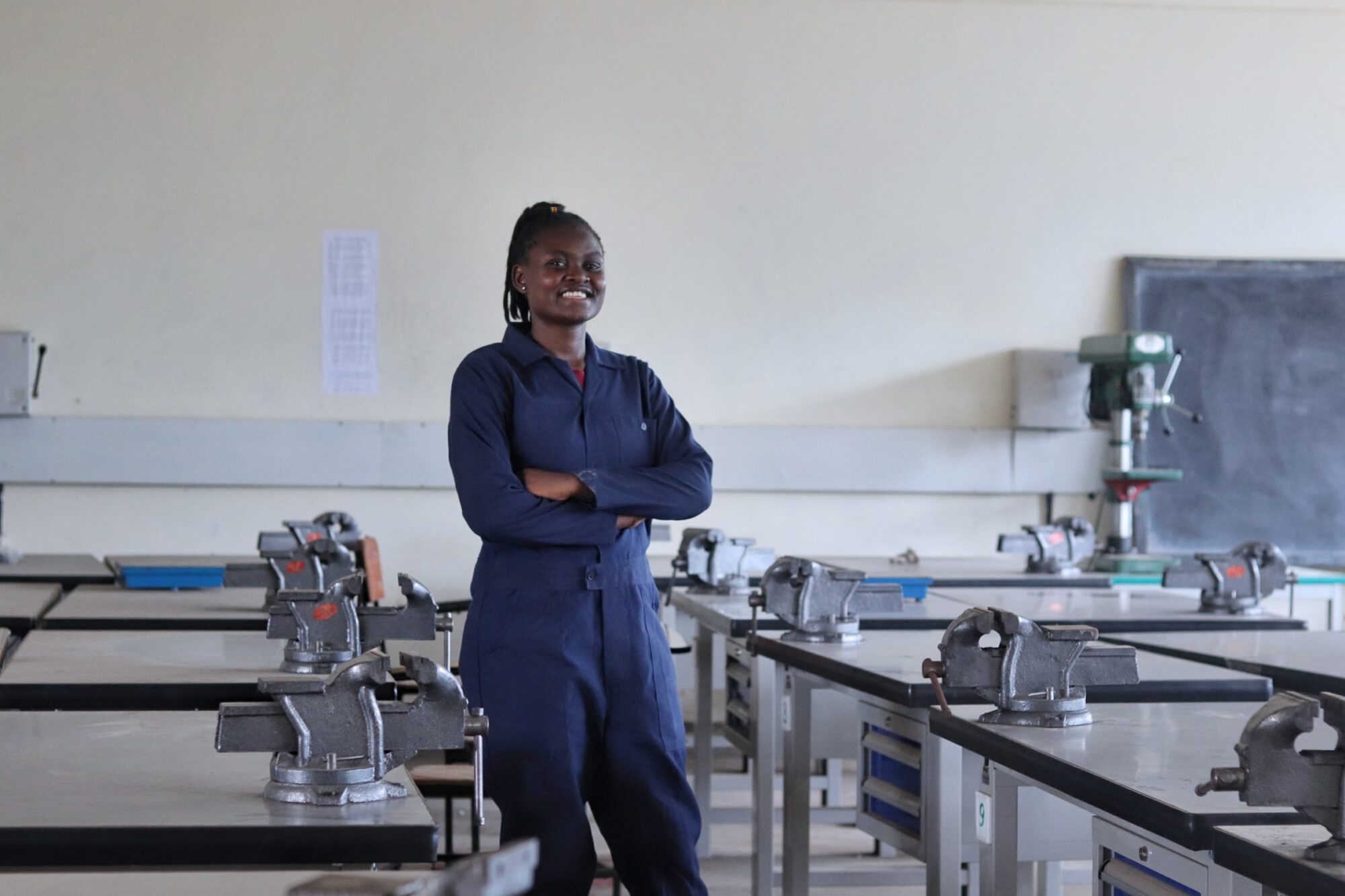 A smiling woman with arms folded wearing a navy jumpsuit stands in a classroom workshop.