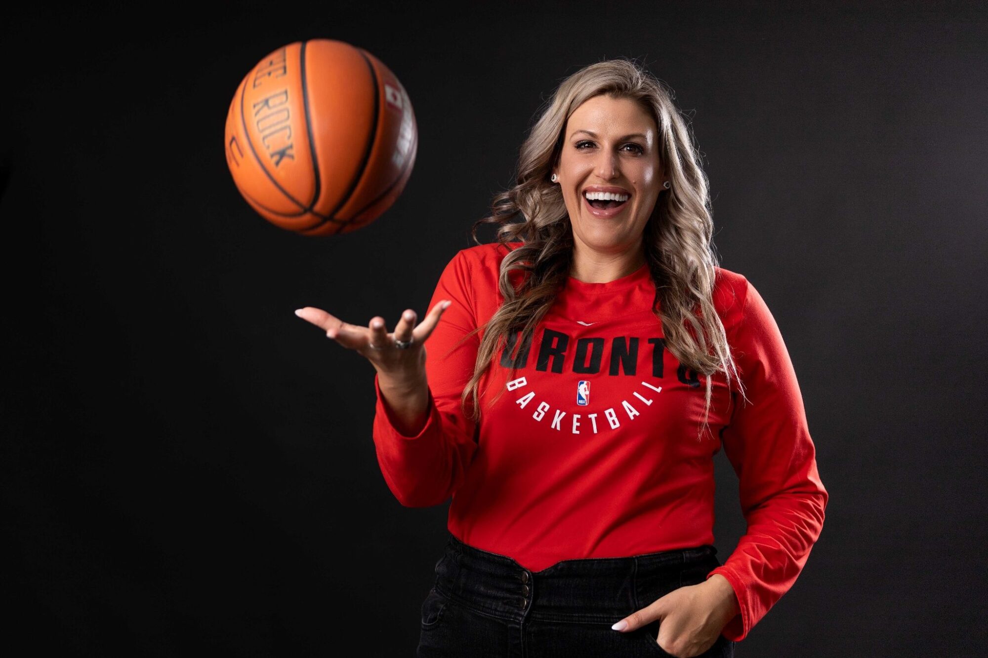 A woman stand smiling at the camera while throwing a basketball in the air