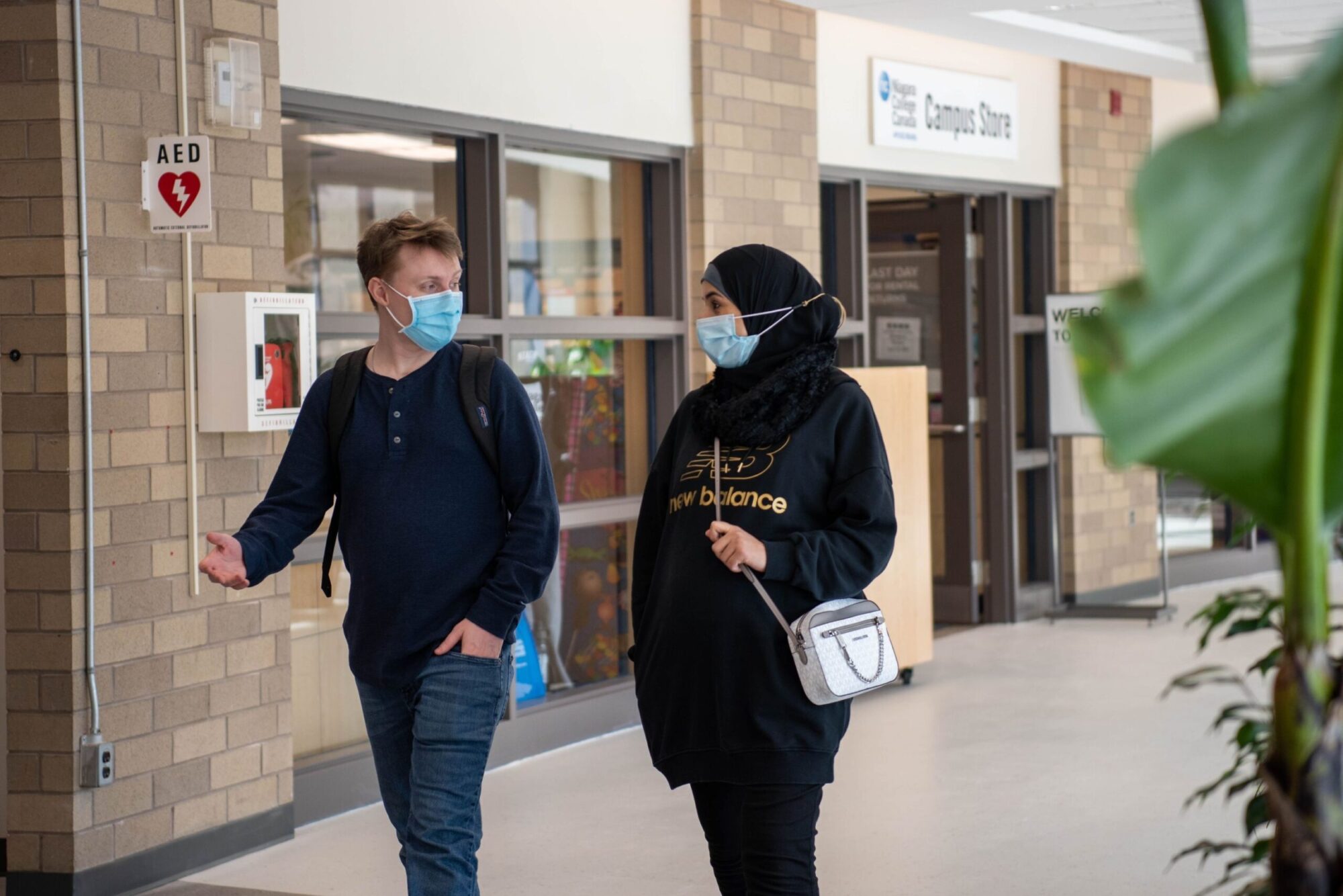 two students wearing medical masks talk while walking down a campus hallway