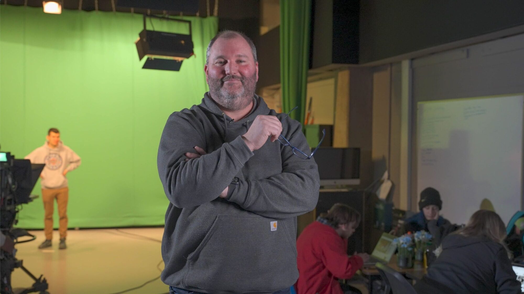 a professor holding eye glasses stands in a television studio on campus with students working in the background