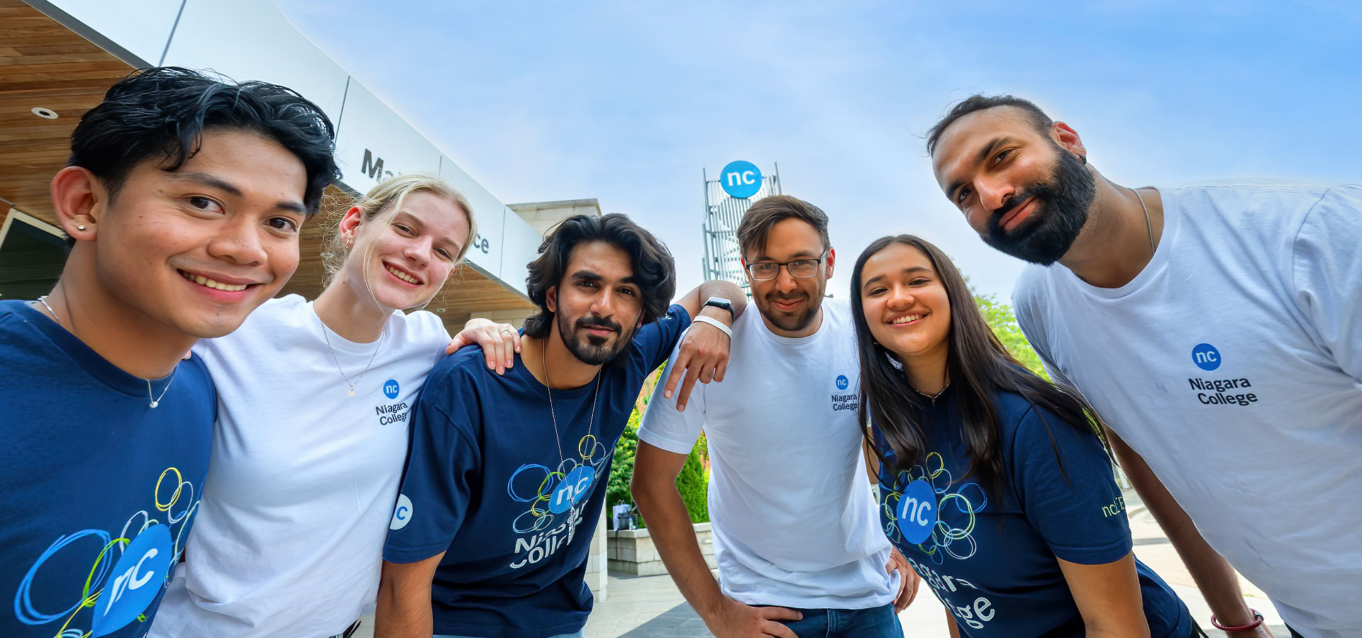 Group of students in NC shirts posing in front of the NC tower at Welland Campus on a bright sunny day