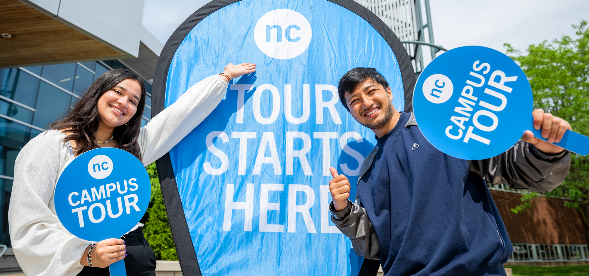 Two NC tour guides holding Campus Tour signs point at a large TOUR STARTS HERE banner