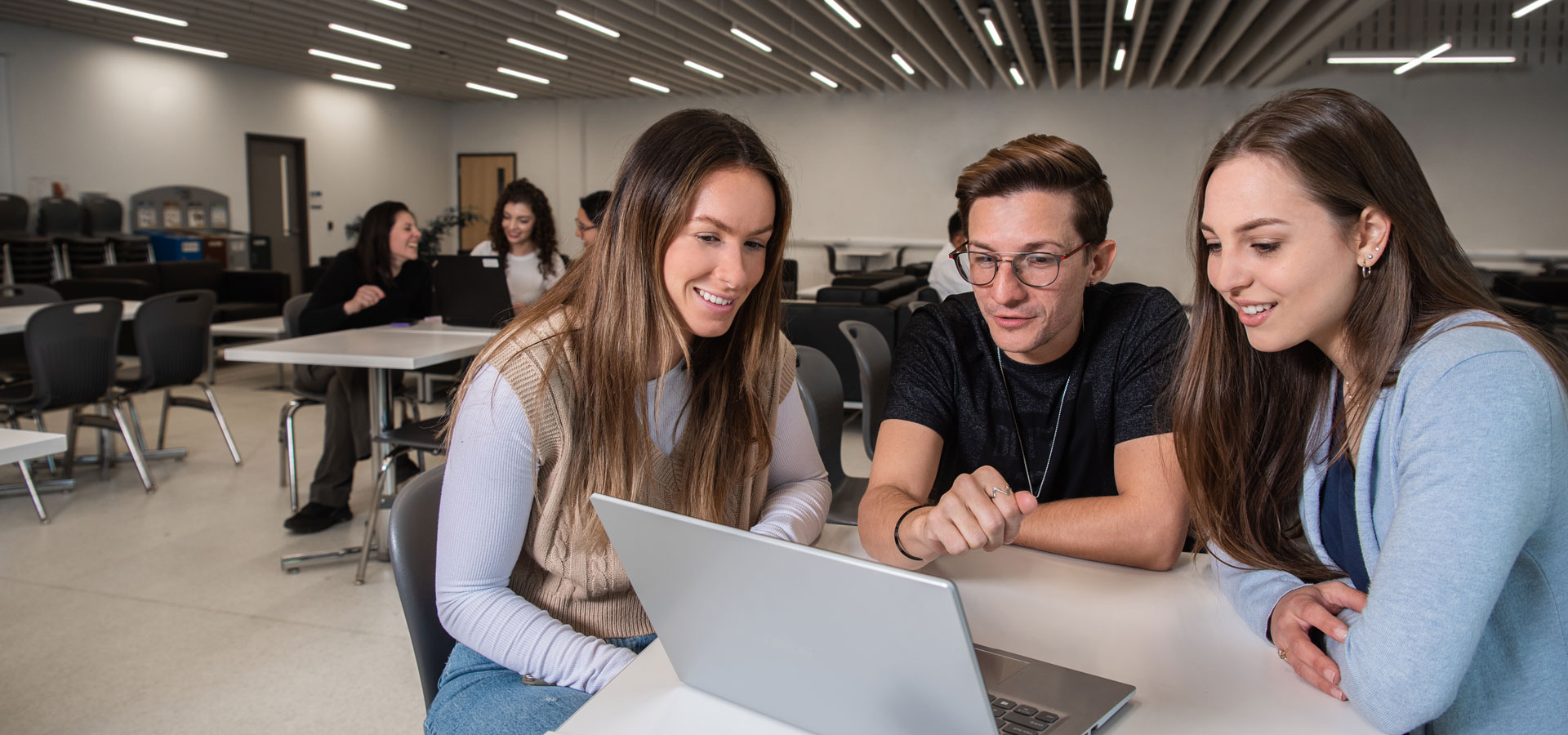 Three students work closely together on a laptop in the modern vaulted space of the NOTL Commons