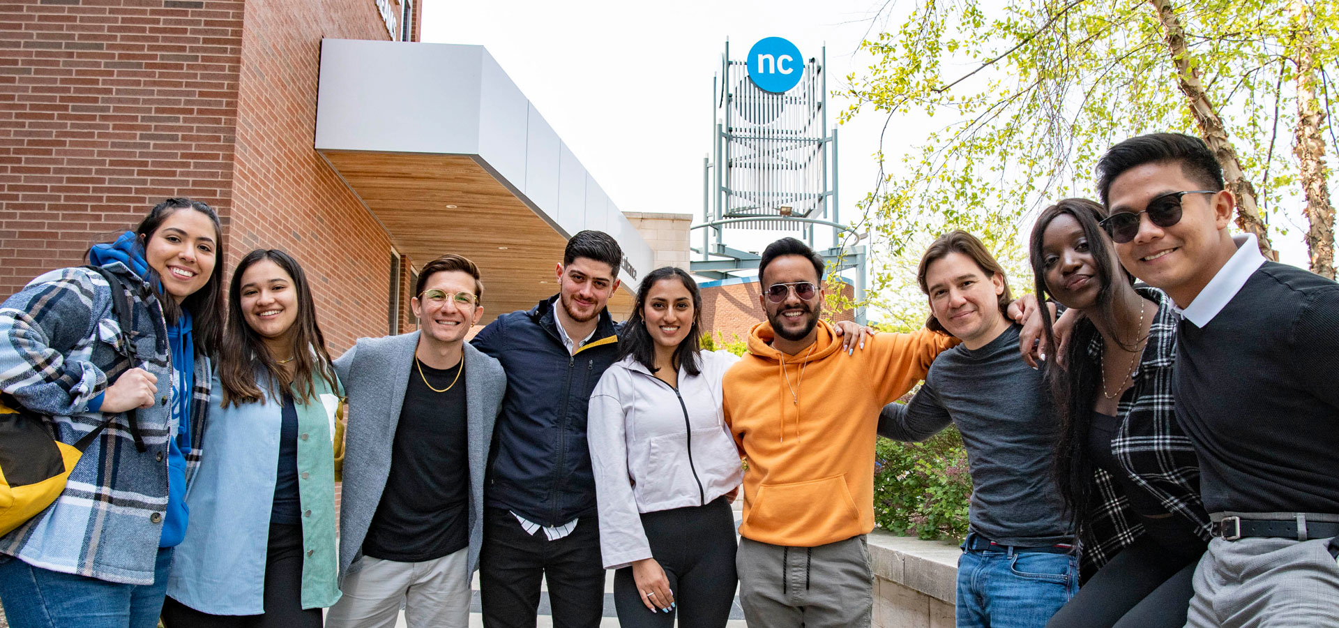 Diverse group of 9 students with their arms over each others shoulders posing in front of the NC tower at Welland campus