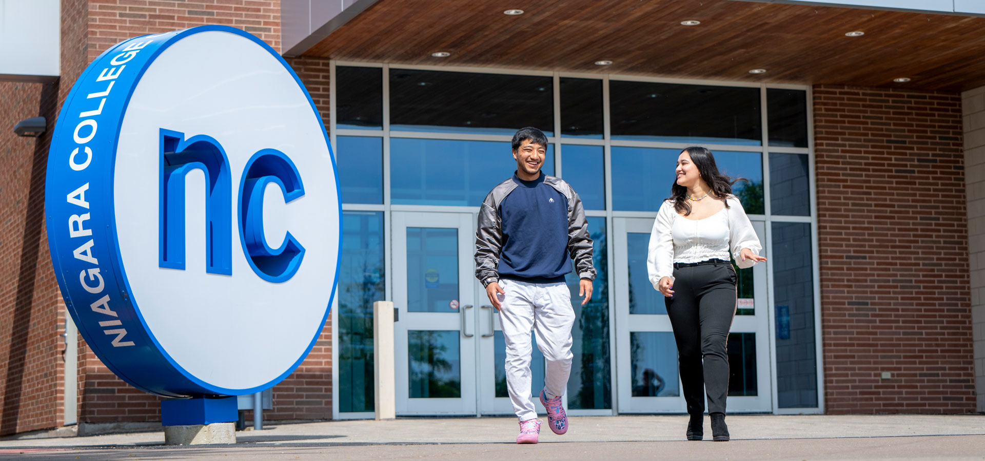 Two students stride out of the front doors next to the big outdoor NC button installation