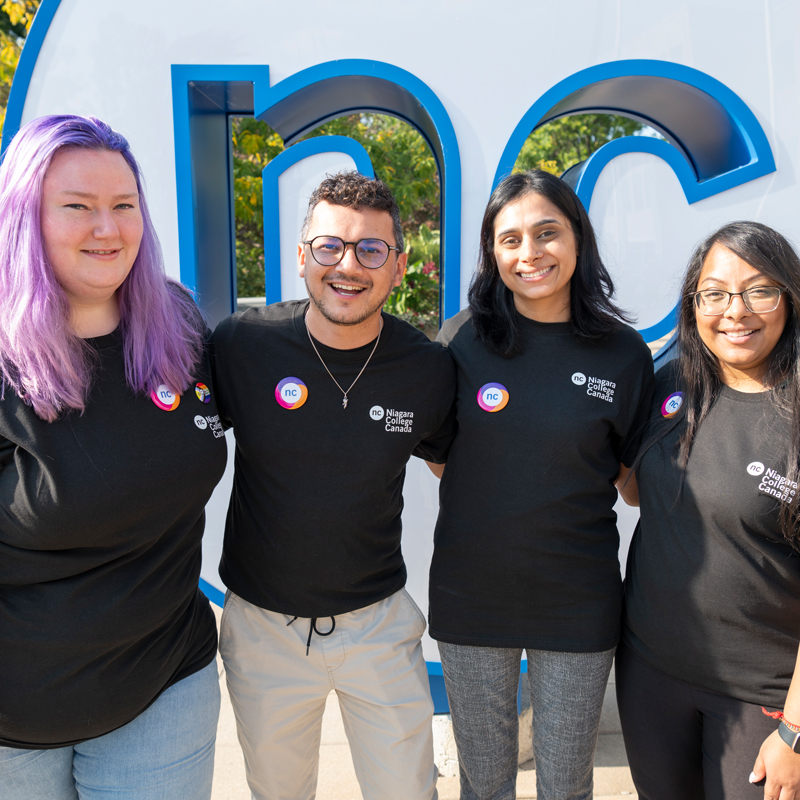 Four students and staff members pose as a group in front of NC sign