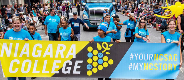 Students wearing NC t-shirts and toques march in the Grape and Wine Parade holding a large NC banner