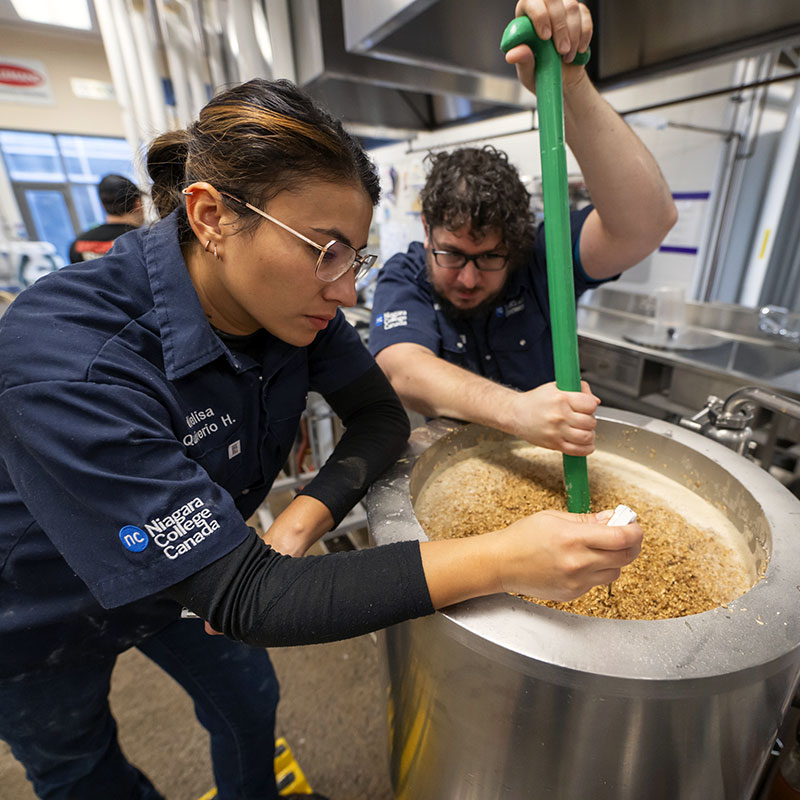 Brewery students wearing branded clothing stir mash in the micro-brewery