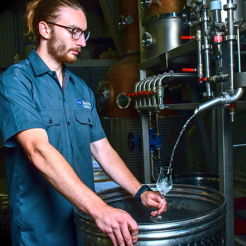 Student distiller in NC distilling uniform tests clear spirits from a spigot