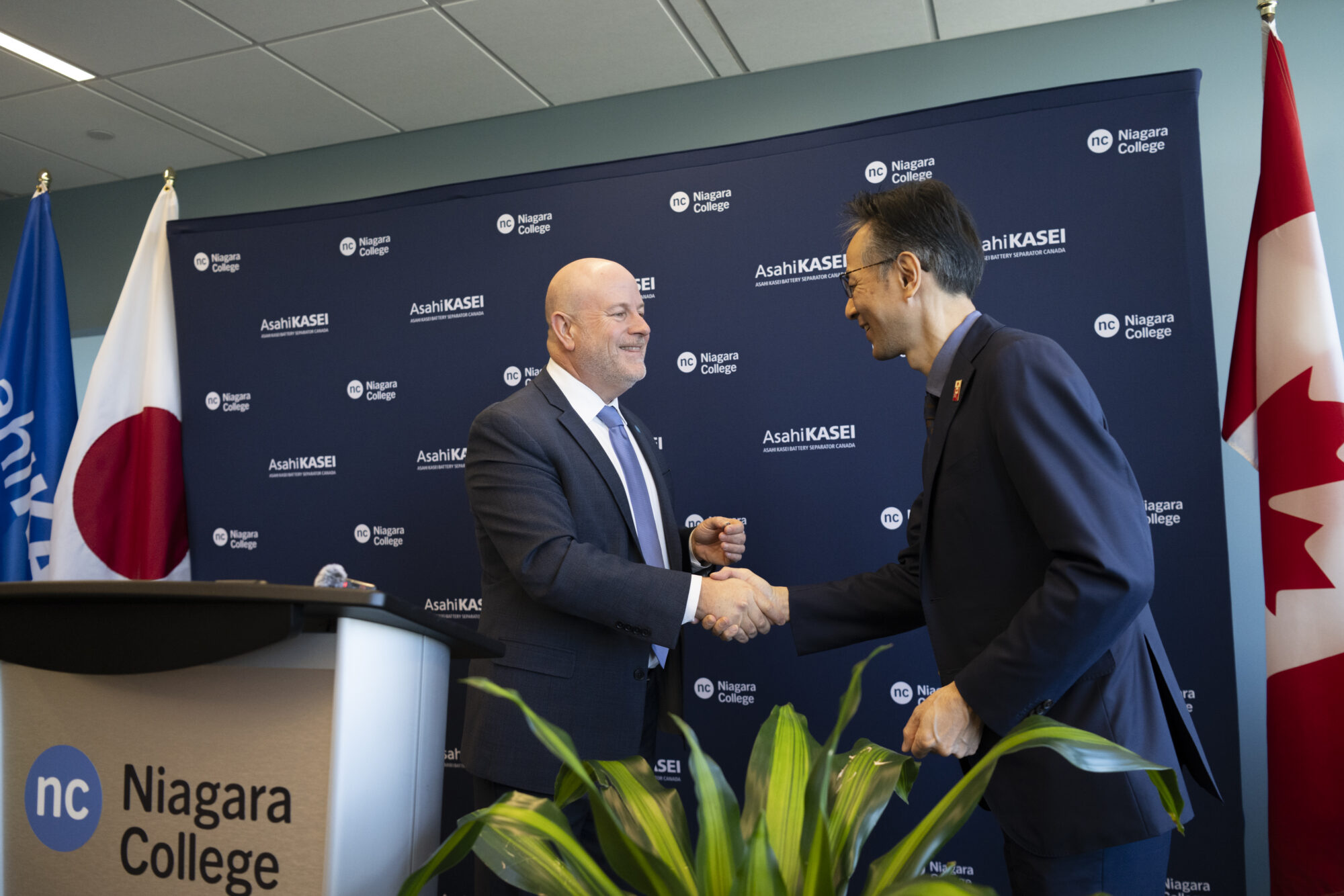 Two men in suits shake hands in front of a backdrop and behind a podium.
