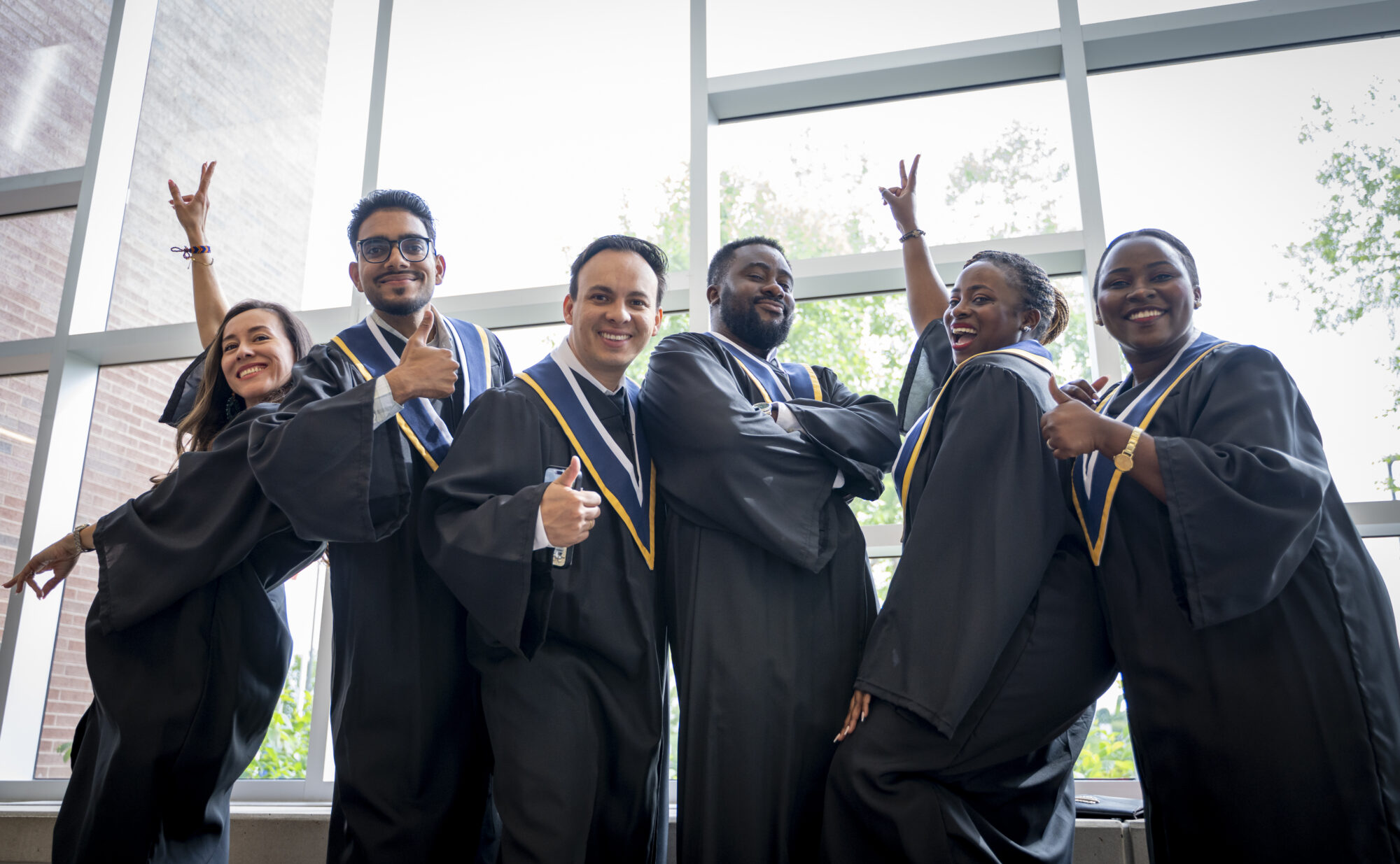 a group of six students wearing black graduation robes pose for a photo, some giving a thumbs up and others raising their arms